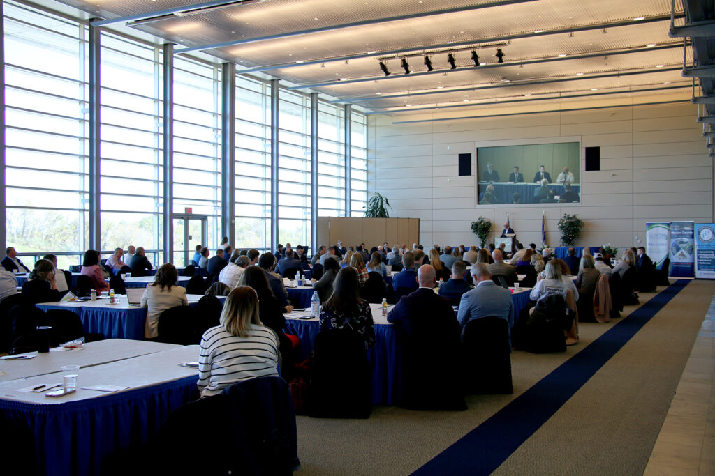 A large audience listens to a panel discussion at the NCWV Business Summit, with the session projected on a screen at the front of the room.