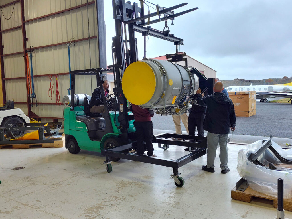 A group of people use a forklift to lift and position a PW306C aircraft engine onto a metal stand inside an aviation hangar.