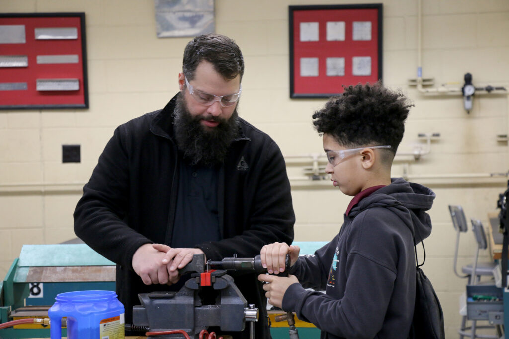 Charles Peters, an instructor in Pierpont’s Aviation Maintenance Technology program, stands beside a student at a workbench and demonstrates how to use a tool secured in a vise inside an aviation lab classroom.