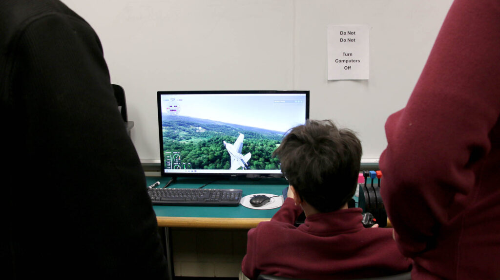A student sits at a computer station operating a flight simulator that displays an aircraft flying over a landscape, with others standing nearby in an aviation lab setting.
