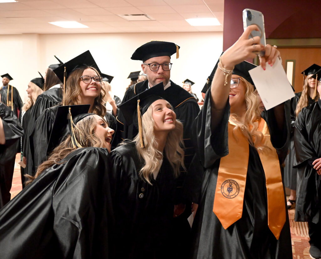 Graduates in caps and gowns take a selfie with a college official before a commencement ceremony, with other graduates gathered in the background.