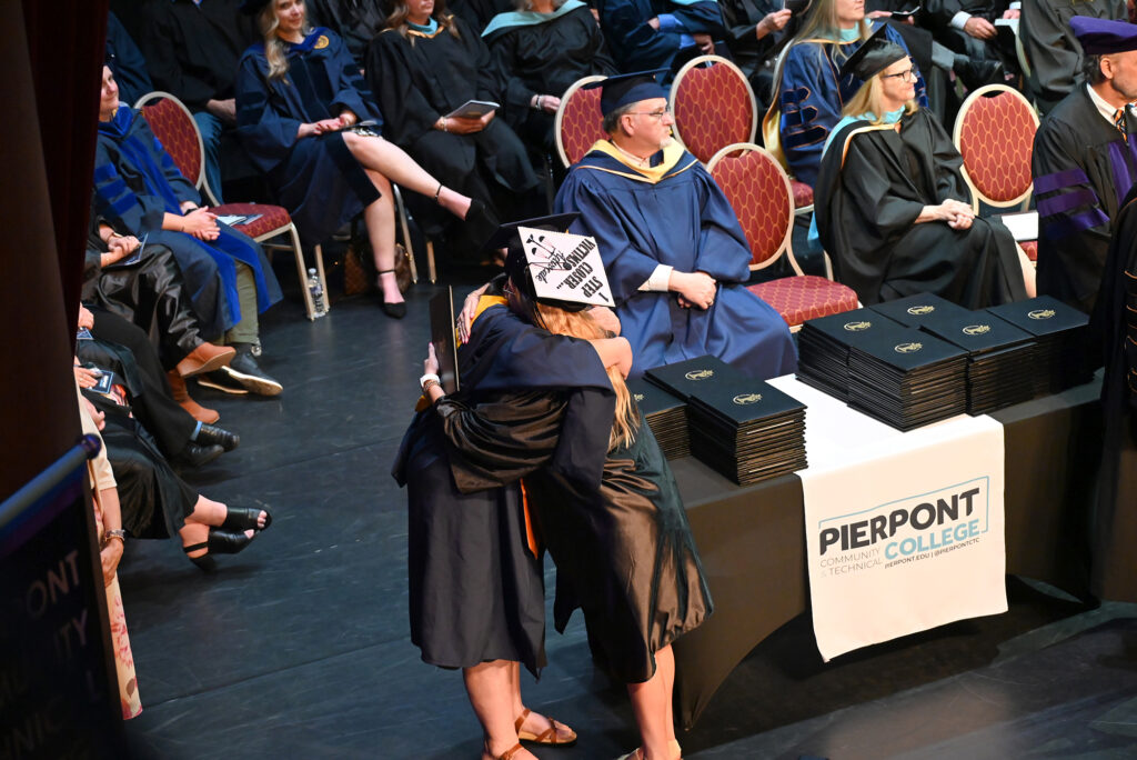 A graduate and college dean in caps and gowns embrace near a table with stacked diploma covers during a commencement ceremony, with faculty seated behind.