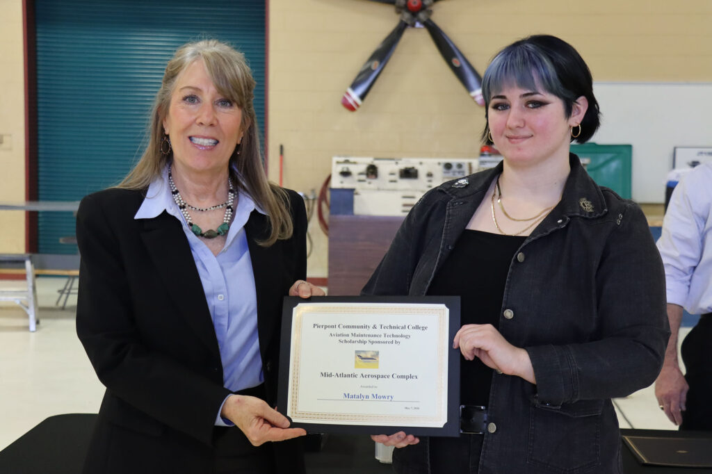 Two people stand in an aviation training space holding a framed scholarship certificate from the Mid-Atlantic Aerospace Complex awarded through Pierpont Community and Technical College’s aviation maintenance technology program.
