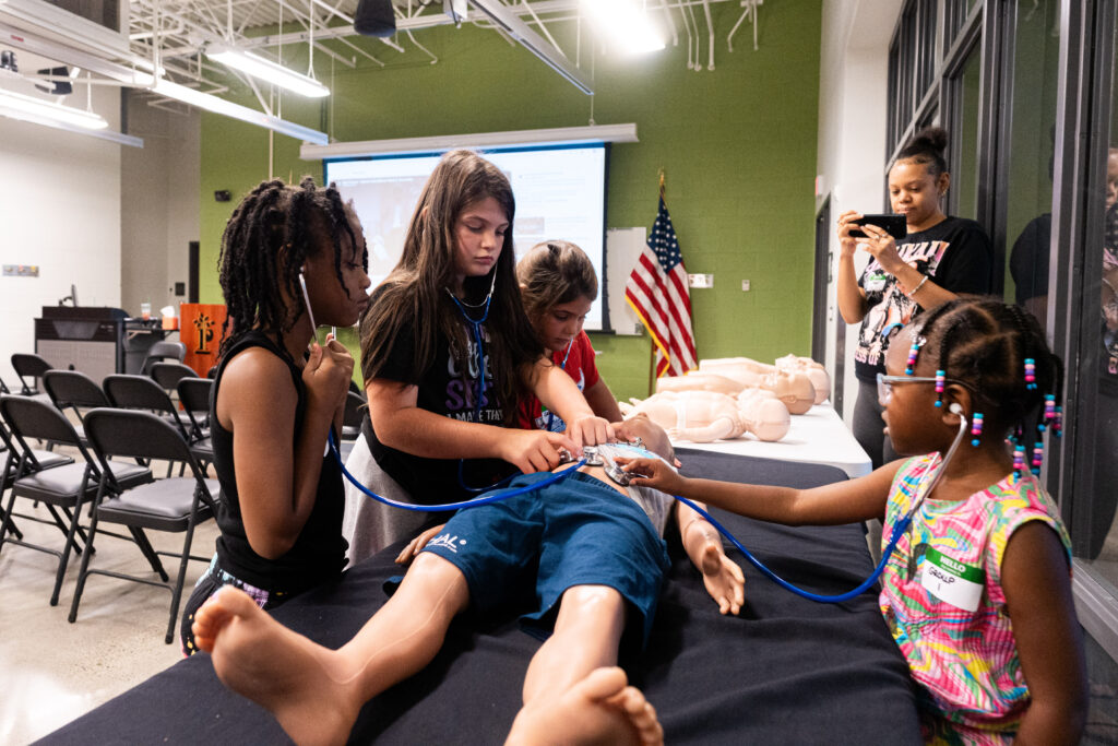 A group of children practice medical skills on a training dummy with stethoscopes, while one person films with a phone.