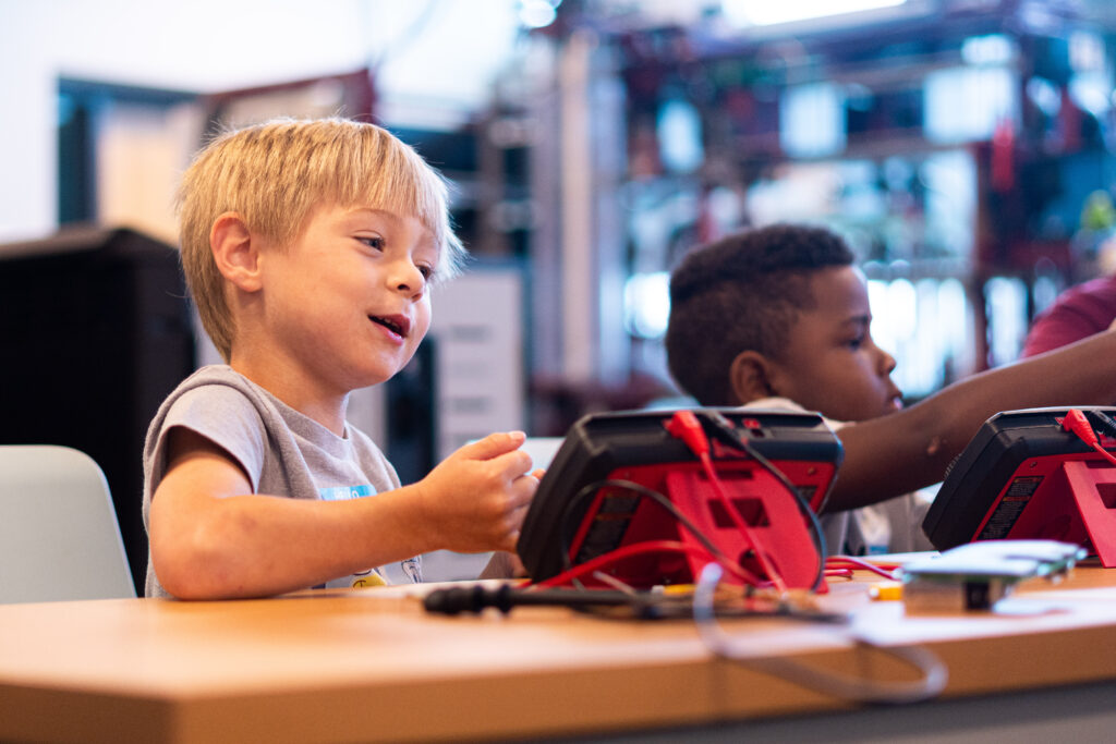 A young child with blond hair looks to the side while sitting at a table with electronic equipment.