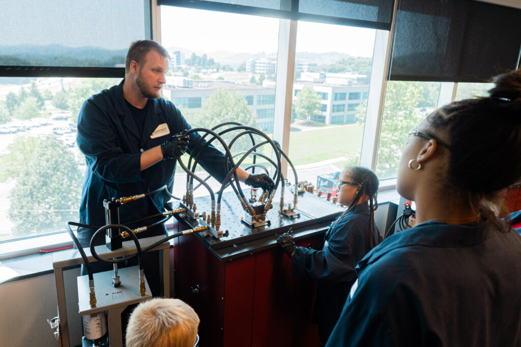A man in a blue lab coat demonstrates a complex hydraulic system to two individuals in a classroom setting, with large windows in the background showing trees and buildings.