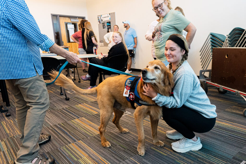 A golden retriever service dog wearing a blue vest is petted by a woman while another person holds its leash. Several other people are visible in the background.