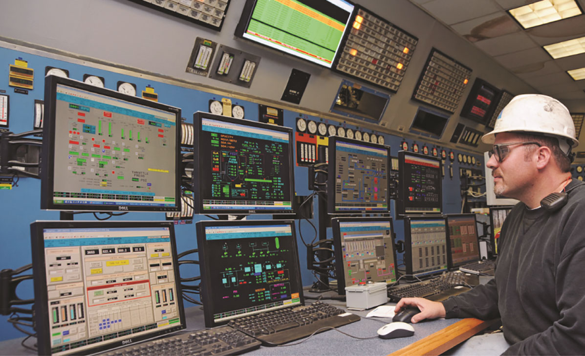 Man wearing hard hat looks at several monitors in a control room.