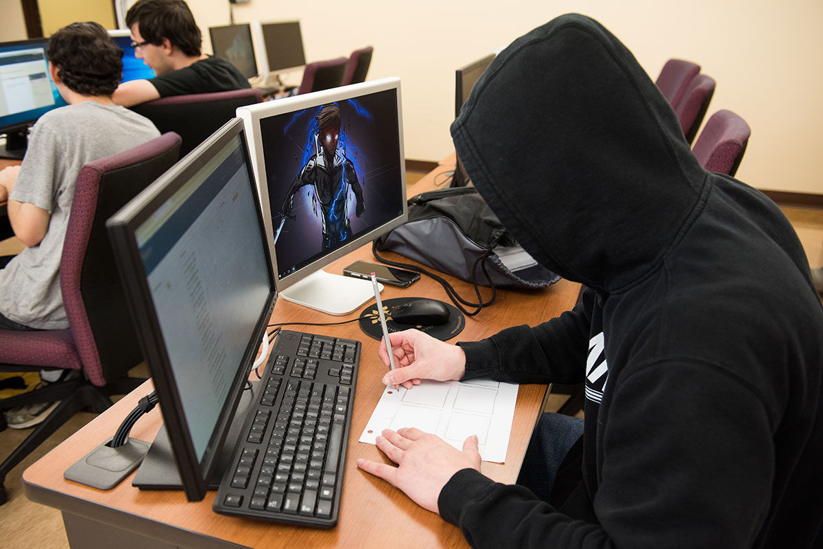 Student sketching storyboard on paper while sitting at a desk with two monitors, a keyboard and mouse.