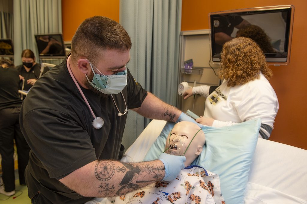 Student places oxygen mask over dummy patient laying in hospital bed