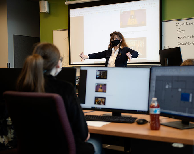 Instructor teaching in classroom as student watches from computer workstation