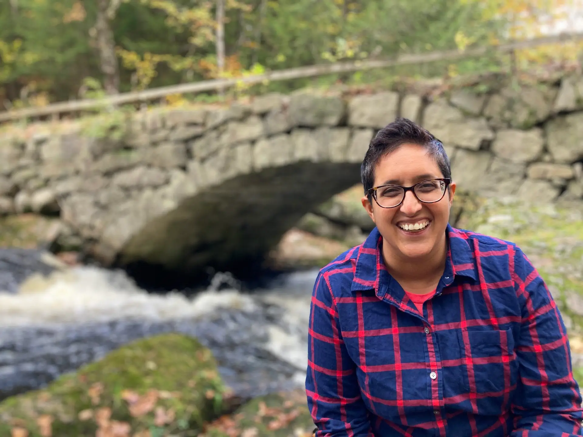 Author Neema Avashia is pictured sitting in front of a wooded area with a stone bridge.