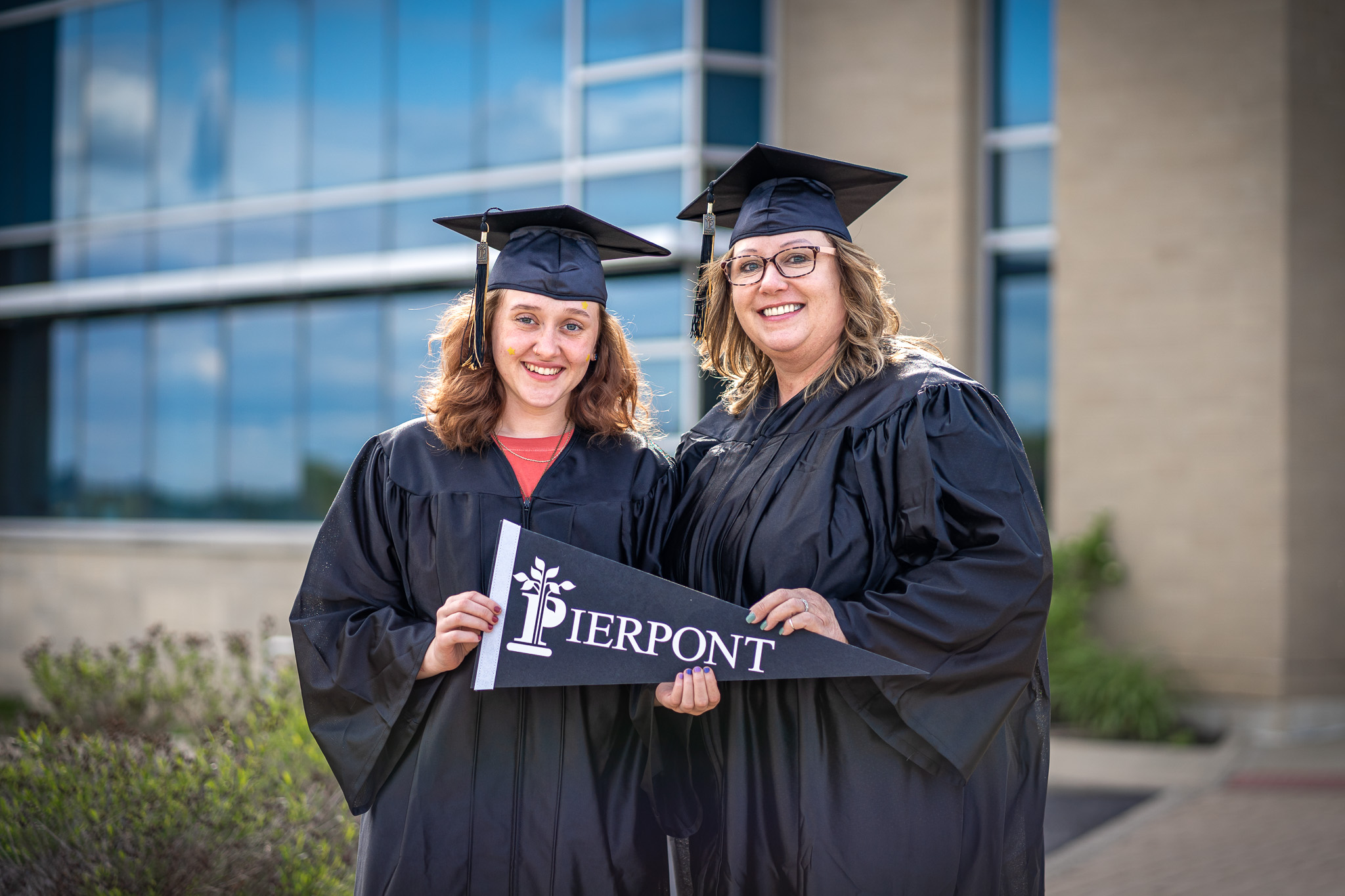 A mother and daughter wearing graduation gowns smile while holding a pennant that reads "Pierpont."