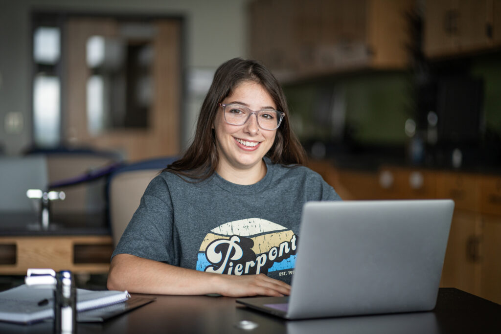 A student smiles in a classroom setting while sitting in front of a laptop.
