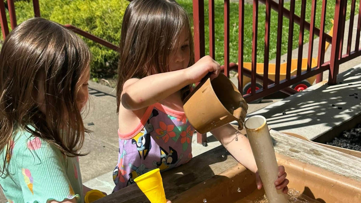 Two children engaged in outdoor water play. One child pours water from a brown container into a vertical white pipe while the other observes. They are next to a wooden water play structure with toys, set against a backdrop of green grass and a red railing.