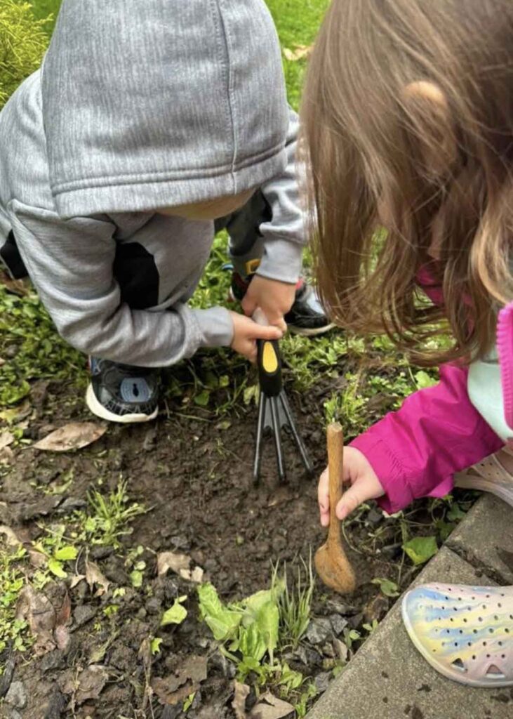 Two young children are gardening together. One uses a hand rake while the other holds a wooden spoon, both focused on digging in the soil.