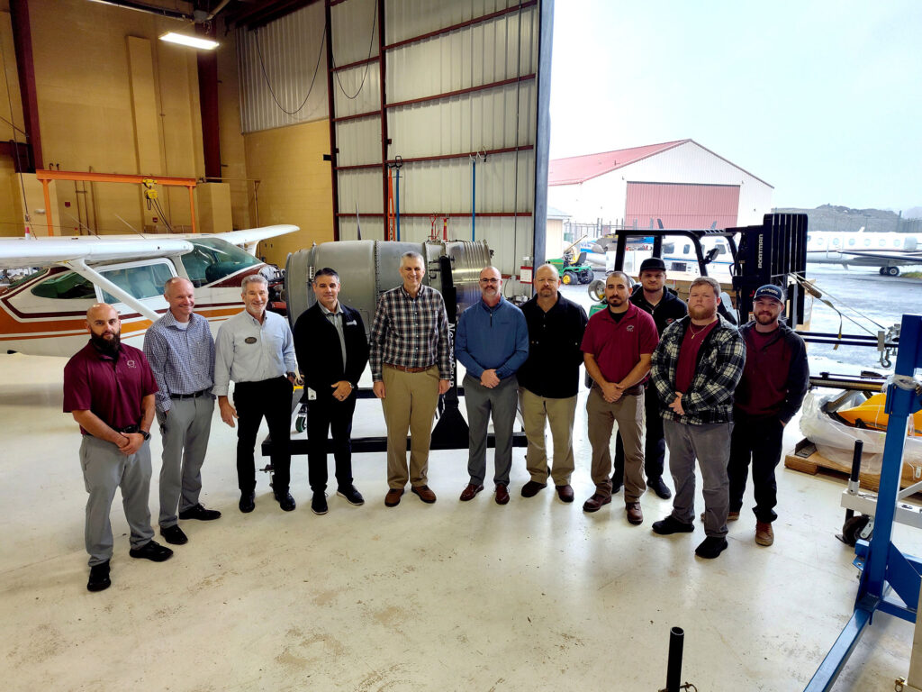 A group of people stand inside an aviation hangar in front of a Pratt & Whitney Canada PW306C engine mounted on a stand, with an aircraft and equipment visible behind them.