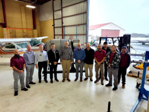 A group of people stand inside an aviation hangar in front of a Pratt & Whitney Canada PW306C engine mounted on a stand, with an aircraft and equipment visible behind them.