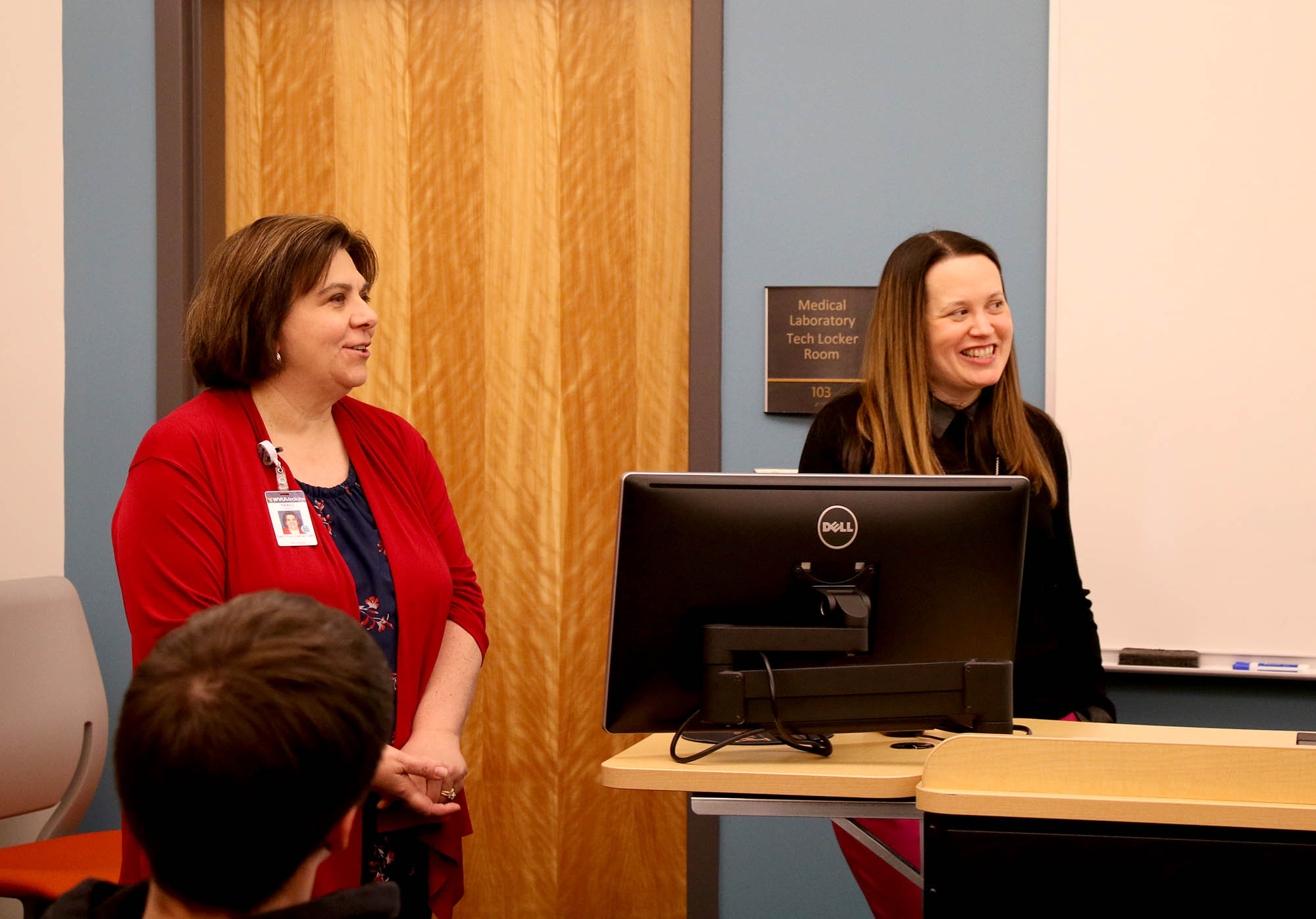Two WVU Medicine representatives stand at the front of a classroom near a computer podium while addressing Medical Laboratory Technology students.