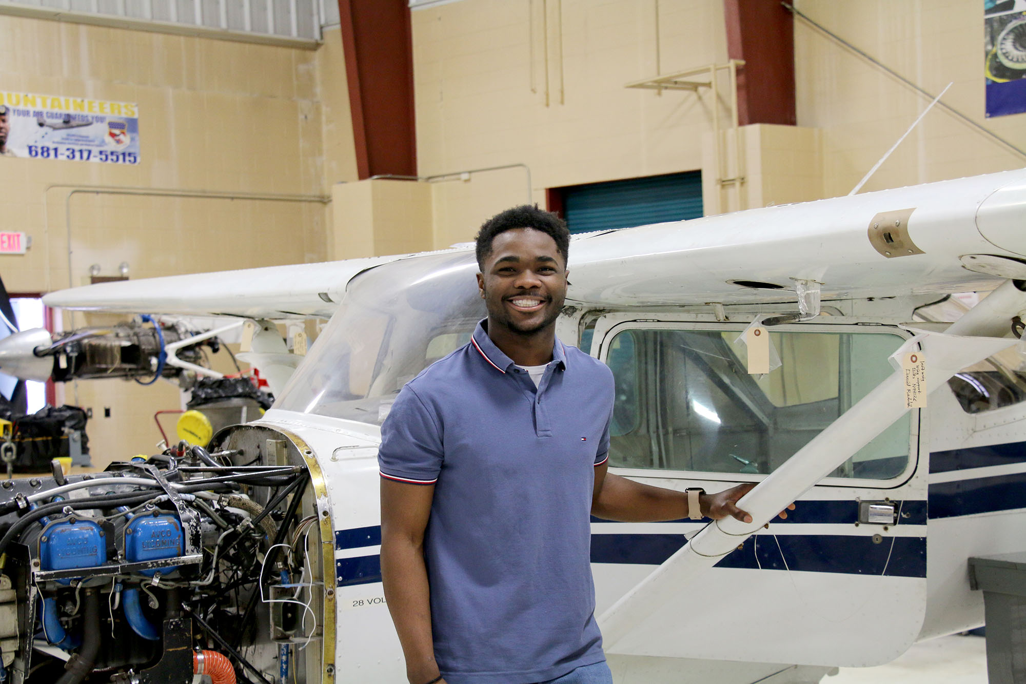 A person wearing a short-sleeve polo shirt stands inside an aircraft hangar at the RCB National Aerospace Center, positioned beside a small white aircraft. An exposed aircraft engine and additional aviation equipment are visible in the background.