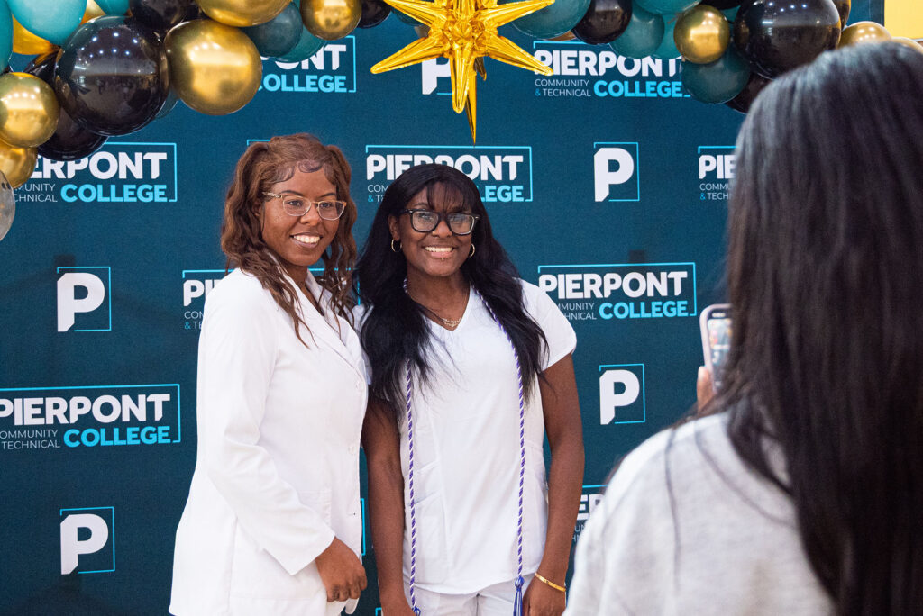 Two graduates in nursing attire pose for a commemorative photo beneath a balloon arch in front of a Pierpont Community & Technical College backdrop during the Licensed Practical Nursing Pinning Ceremony, while an attendee takes their picture.